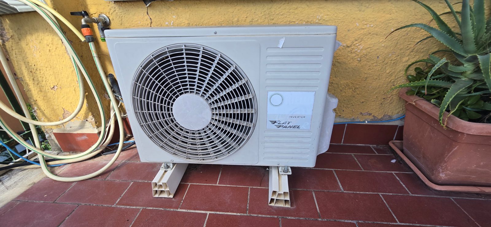 Older white mini-split outdoor unit with INVERTER and FLAT PANEL markings, standing on two floor feet on a red tiled terrace, its side panel close to a yellow rendered wall, with coiled garden hoses on the left and a potted aloe on the right.